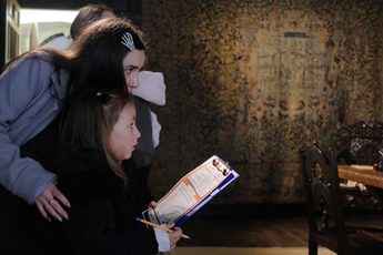 An adult and child side view looking at something out of the frame. The child holds a pencil and clipboard.