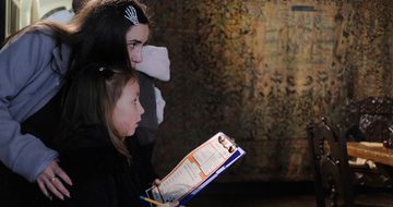 An adult and child side view looking at something out of the frame. The child holds a pencil and clipboard.