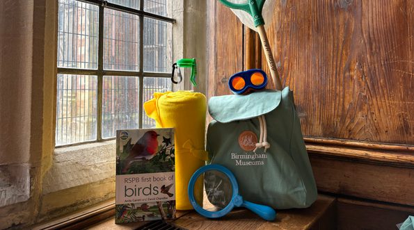A window ledge with an explorer backpack and items including a net, magnifying glass, binoculars, book and blanket.