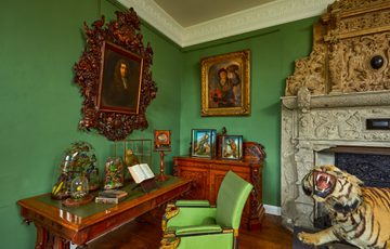 A green groom with desk and chair. Taxidermy birds are in cases on the desk and taxidermy tiger to the side.