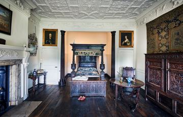 A bedroom with wooden four-poster bed, ornate ceiling, fireplace with paintings on the wall.