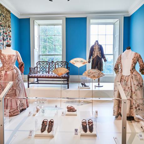Three historical costumes and shoes on display with tapestry on the wall.