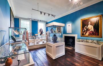 Blue room with display cases featuring ceramic objects, paintings on the wall and costumes on display.
