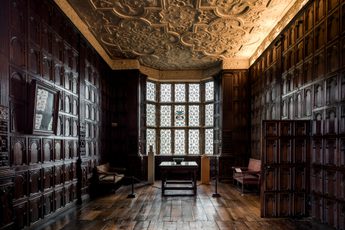 Dark wooden panel walls and wooden floors are lit by the light of a window.