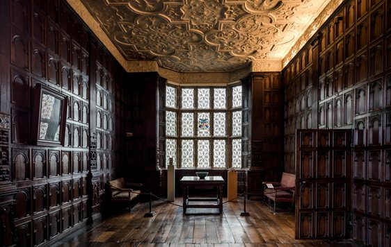 Dark wooden panel walls and wooden floors are lit by the light of a window.