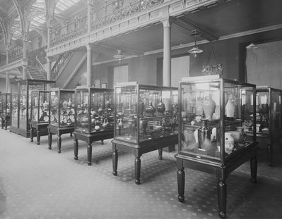 Black and white photo of part of the Industrial Gallery with rows of glasses cases containing objects.