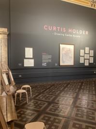 An gallery space with dark walls, drawings on display and easels for people to use.