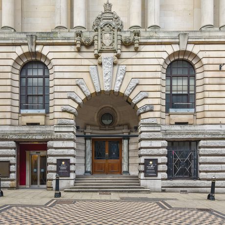 Historic stone building with arched windows, grand entrance, decorative sculptures, and checkered pavement.