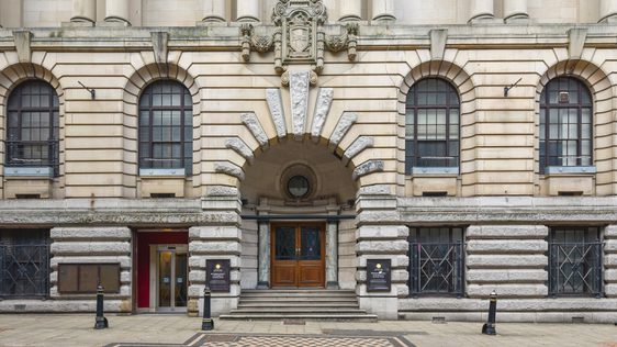 Historic stone building with arched windows, grand entrance, decorative sculptures, and checkered pavement.