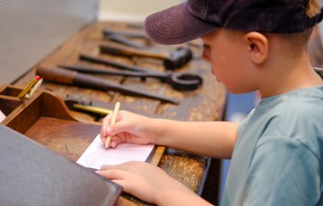 A boy wearing a cap writes on paper while sitting at a wooden workbench.
