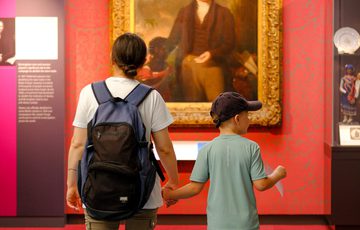 A person and child hold hands while viewing a large, framed portrait in a museum.