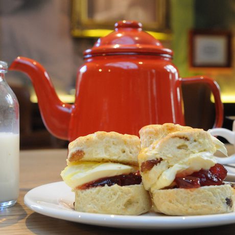 Scones with cream and jam, red teapot, bottle of milk, and white teacup on table.