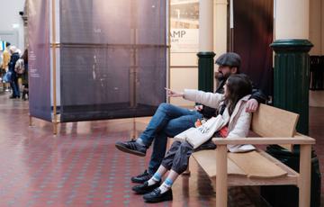 An adult and child sitting relaxing on a bench in a gallery. The child is pointing at something out of shot.