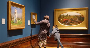 An adult and child looking at paintings in gold frames on display in an art gallery.