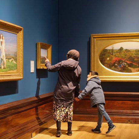 An adult and child looking at paintings in gold frames on display in an art gallery.