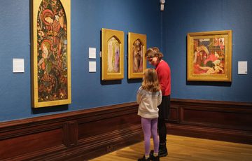 An adult and child looking at paintings in gold frames on display in an art gallery.