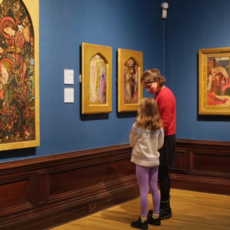 An adult and child looking at paintings in gold frames on display in an art gallery.