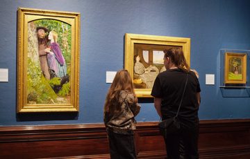 An adult and child looking at paintings in gold frames on display in an art gallery.