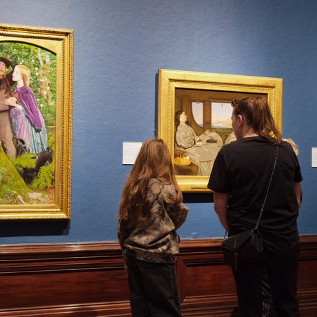 An adult and child looking at paintings in gold frames on display in an art gallery.
