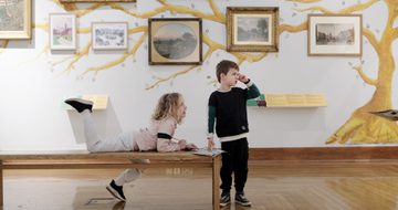 Two children, one is sitting on a bench, looking into a gallery, artwork is on display behind them.