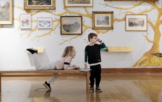 Two children, one is sitting on a bench, looking into a gallery, artwork is on display behind them.