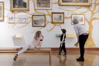 An adult and two children, one who is sat on a bench, are looking at paintings on display.