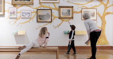 An adult and two children, one who is sat on a bench, are looking at paintings on display.