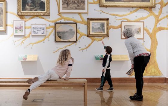 An adult and two children, one who is sat on a bench, are looking at paintings on display.