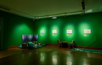 A gallery display with artwork on green walls, a small soft play area with a table and chairs.