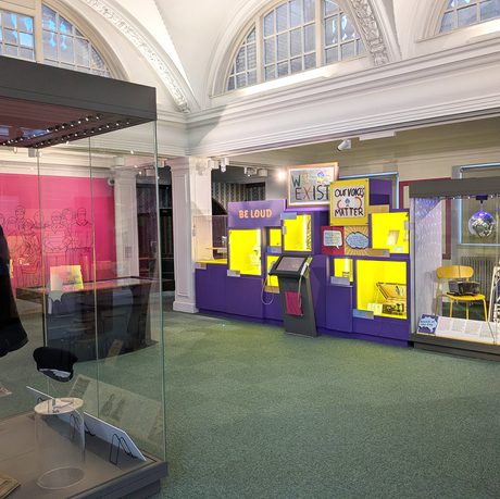 A pink and purple gallery space featuring large glass display cases with costume, furniture posters and other objects.