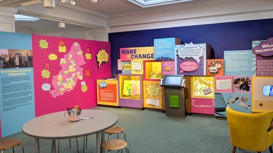 Colourful gallery space fearing chairs table, display cases and speech bubble graphics.