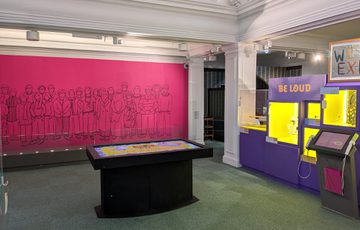 Gallery display featuring touch table, mural, and purple display cases with the text 'Be Loud'.