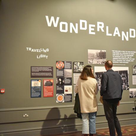 Two people standing in front of a wall of information and images, on the wall is the in larger lettering it says 'Wonderand'
