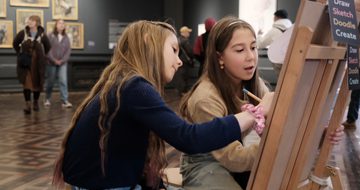 Close up of two children sitting and drawing at an easel in a gallery.