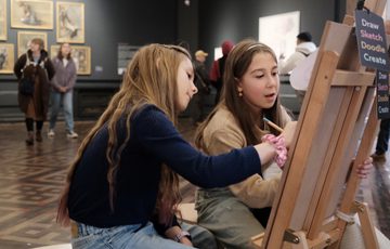 Close up of two children sitting and drawing at an easel in a gallery.