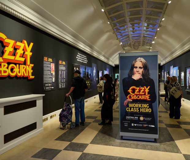 Ozzy Osbourne exhibition with large 'Ozzy Osbourne' graphic, black walls and people in the exhibition space.