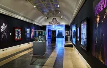 Ozzy Osbourne exhibition with black walls, large photos on display and display cabinet of awards in the centre.