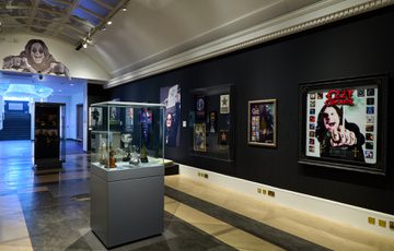 Ozzy Osbourne exhibition with black walls, large photos on display and display cabinet of awards in the centre.
