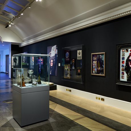 Ozzy Osbourne exhibition with black walls, large photos on display and display cabinet of awards in the centre.
