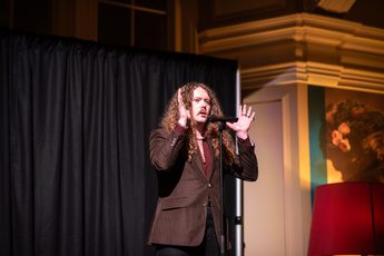 Bradley Taylor performing on stage with microphone and dramatic lighting, his hands are to the up to the side of his head.