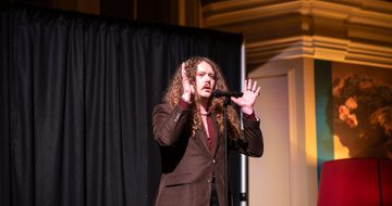 Bradley Taylor performing on stage with microphone and dramatic lighting, his hands are to the up to the side of his head.