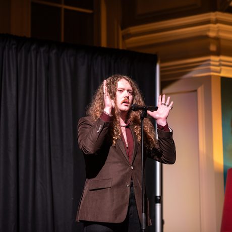 Bradley Taylor performing on stage with microphone and dramatic lighting, his hands are to the up to the side of his head.