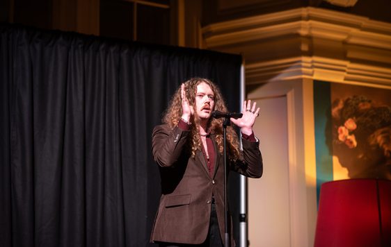 Bradley Taylor performing on stage with microphone and dramatic lighting, his hands are to the up to the side of his head.