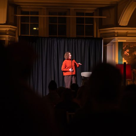 A person performs on stage with microphone, audience watches in dimly lit venue.