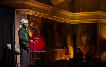 A performer speaks into a microphone on stage, in the background are large portraits and warm lighting.