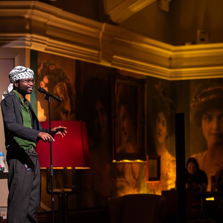 A performer speaks into a microphone on stage, in the background are large portraits and warm lighting.