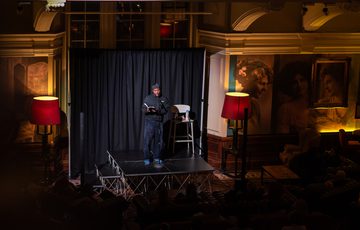 A performer on stage with a microphone stands in a warmly lit room with the audience in darkness.