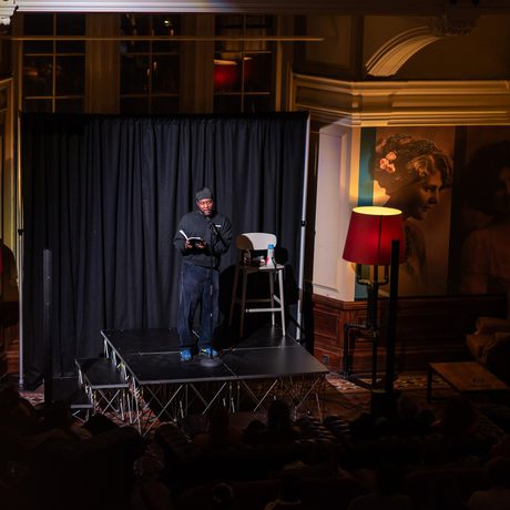 A performer on stage with a microphone stands in a warmly lit room with the audience in darkness.
