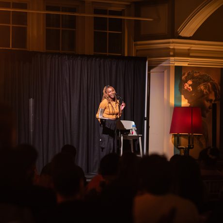 A woman performs on stage lit by warm light, the audience are dark silhouettes.