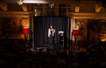A woman performs stand-up comedy on stage lit by warm lighting while the audience are in darkness.